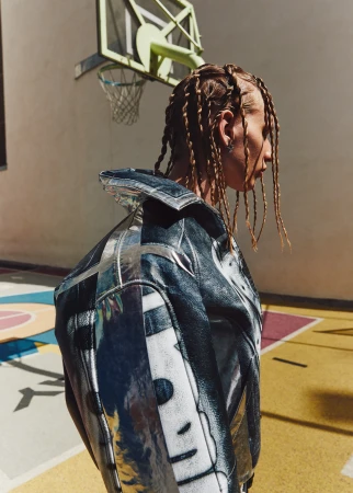 Profile view of a person with blonde braids wearing a shiny silver jacket stands on a yellow and red outdoor basketball court.