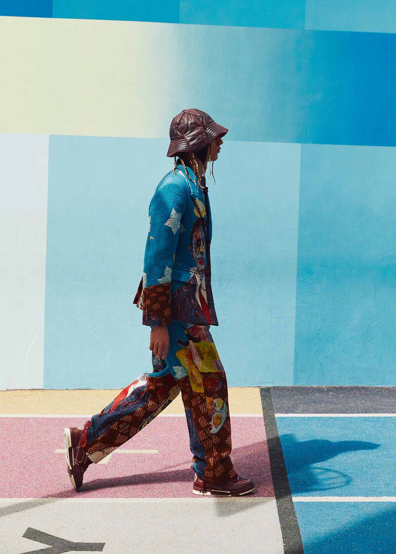 Person walking, wearing patterned blue blazer, pants, brown bucket hat and shoes, on a blue and pink surface, by a colorful wall.