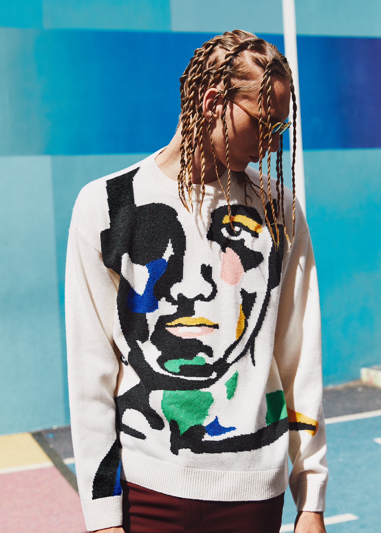 Young man wearing an art-inspired sweater stands against a colorful geometric wall, showcasing modern fashion with a creative edge.
