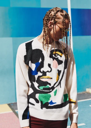Young man wearing an art-inspired sweater stands against a colorful geometric wall, showcasing modern fashion with a creative edge.