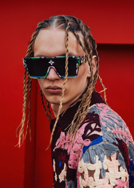 Portrait of a young adult male wearing designer sunglasses and a colorful sweater against a bright red background in a studio shot.