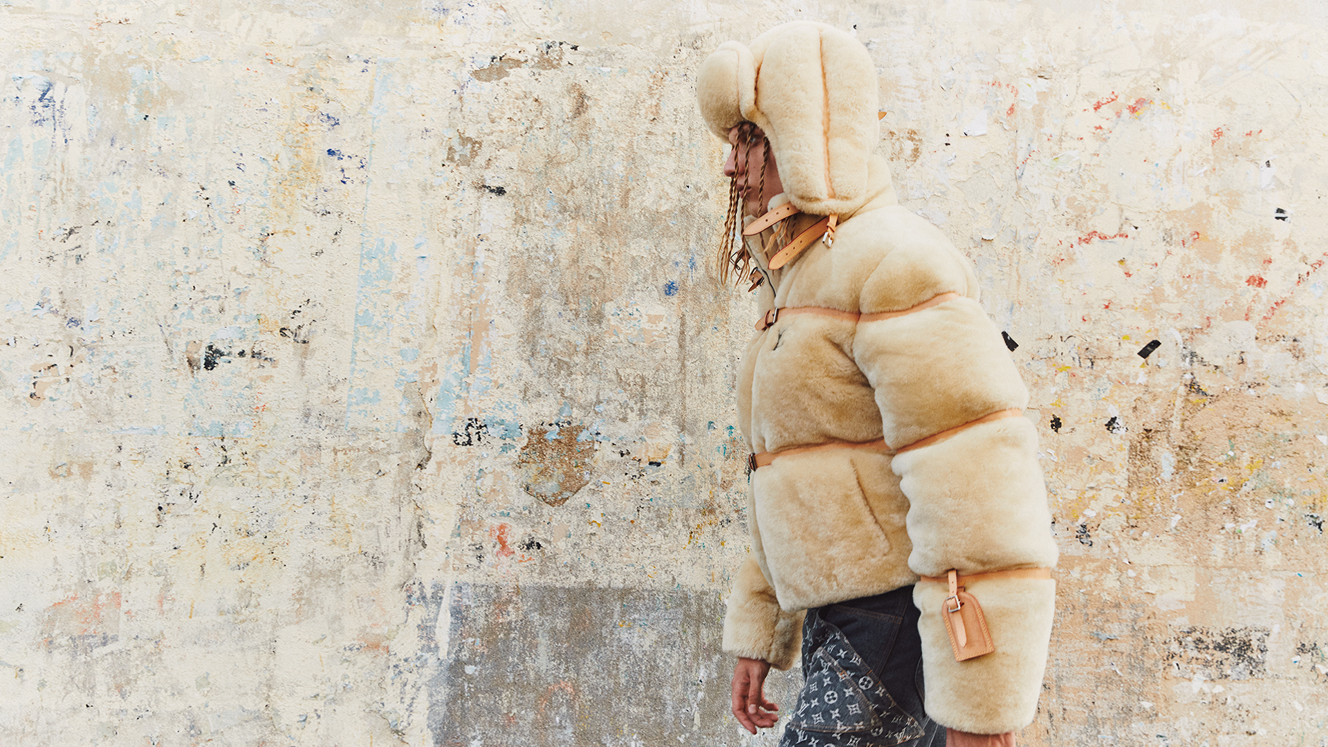 Person in a cream shearling puffer jacket and hat against a textured wall, showcasing a fashion-forward winter look with visible brand logos.