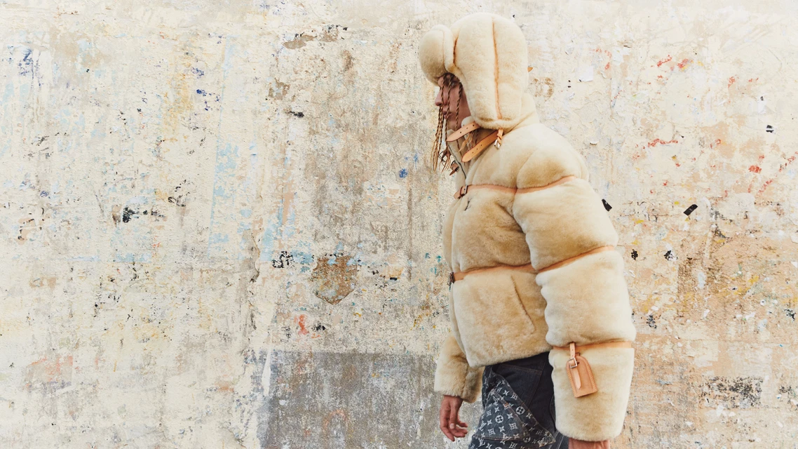 Person in a cream shearling puffer jacket and hat against a textured wall, showcasing a fashion-forward winter look with visible brand logos.
