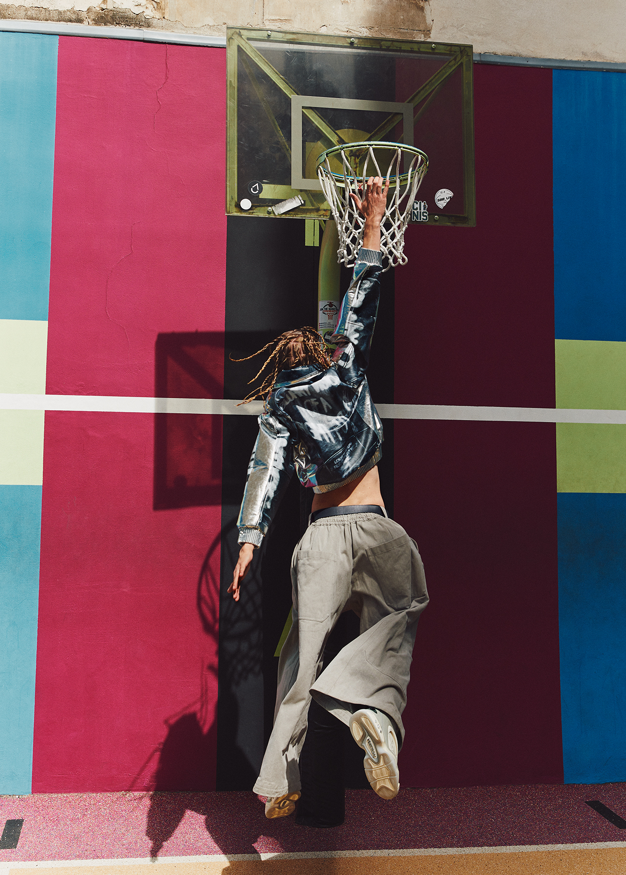 Back view of a person with dreadlocks jumping, attempting to grab a basketball hoop in front of a colorful wall on a sunny day.