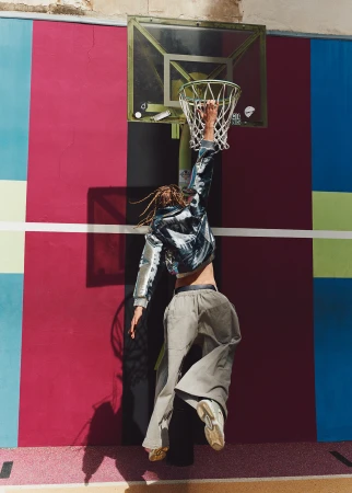 Back view of a person with dreadlocks jumping, attempting to grab a basketball hoop in front of a colorful wall on a sunny day.