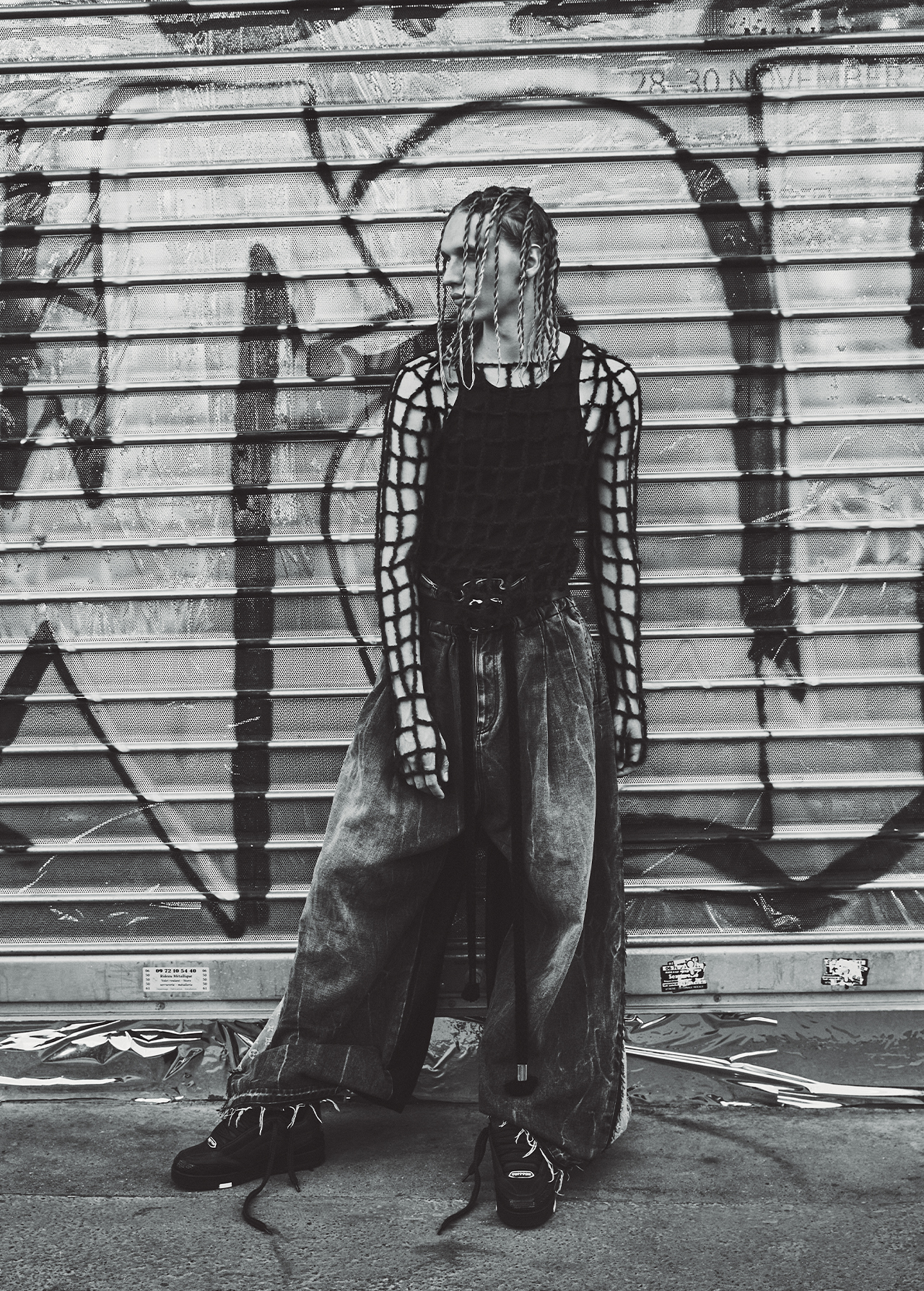Fashionable young adult in a black tank top and baggy jeans, standing against a weathered metal wall with graffiti in black and white.
