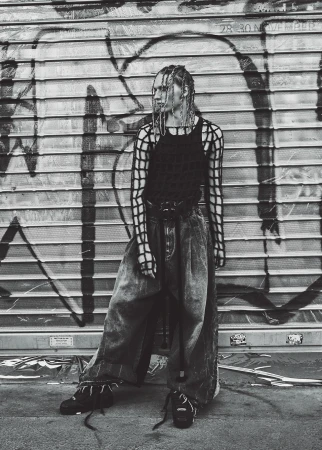 Fashionable young adult in a black tank top and baggy jeans, standing against a weathered metal wall with graffiti in black and white.