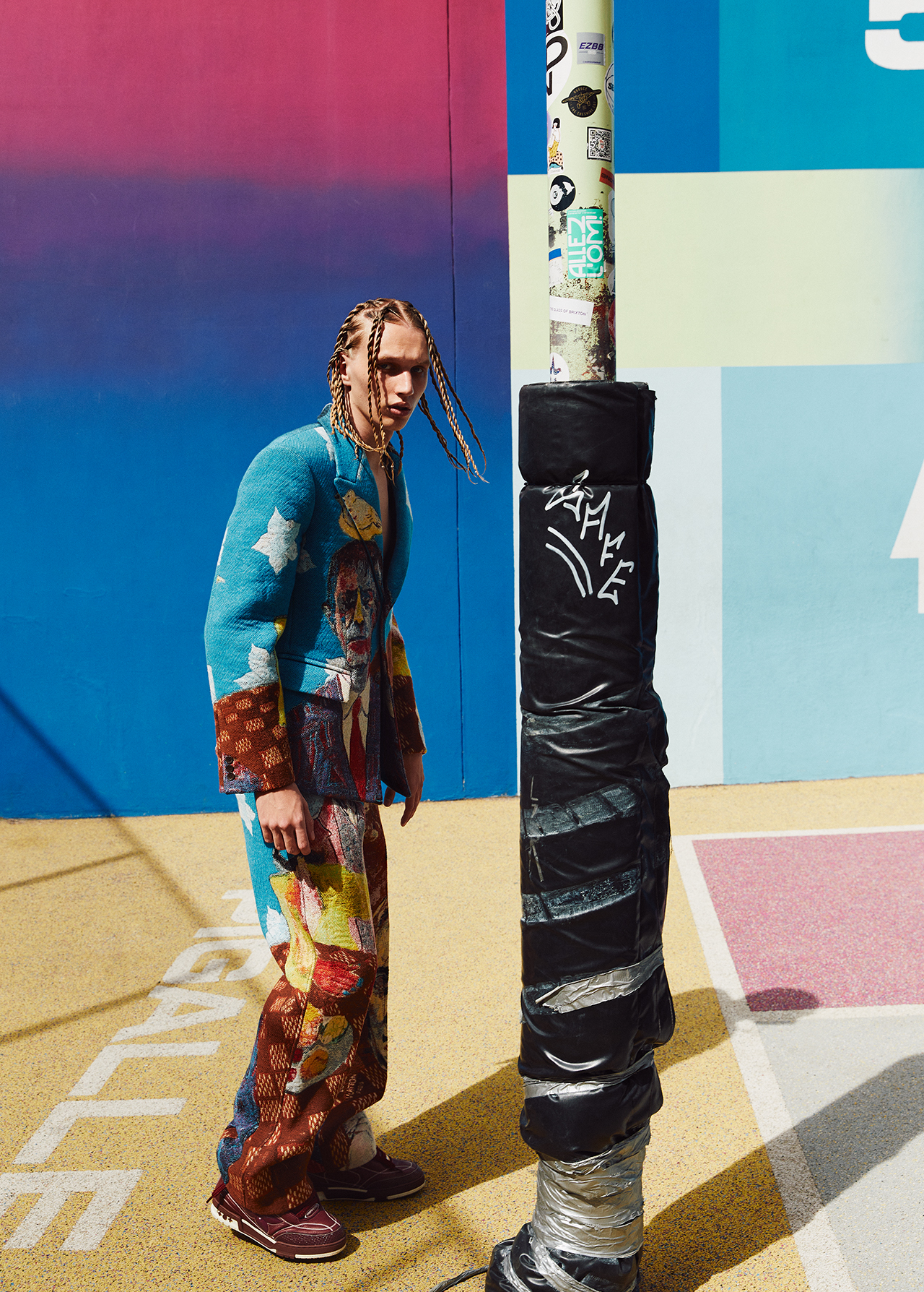 Adult male fashion model in a colorful patterned suit and braids poses outdoors near a painted wall, creating a vibrant editorial image.
