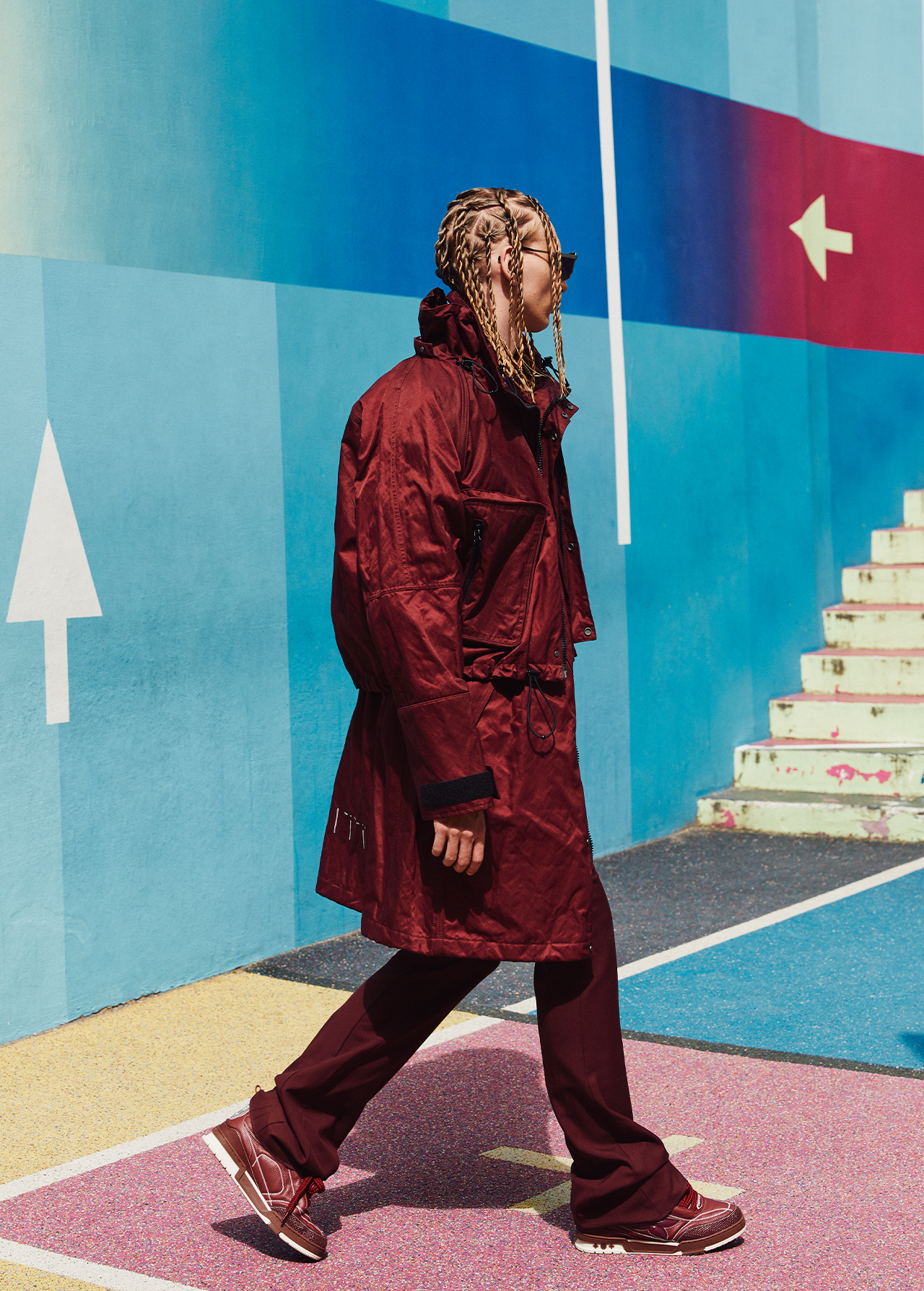 Person walking in a maroon jacket and pants against a painted geometric wall on a sunny day, showcasing streetwear fashion.