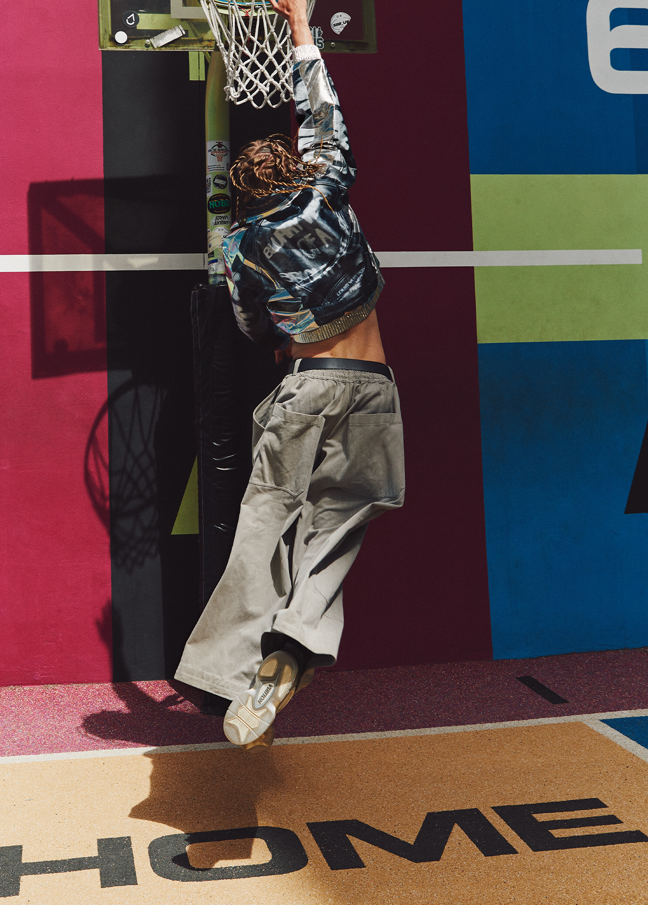 An athletic adult performs a slam dunk on an outdoor basketball court, wearing a metallic jacket and khaki pants in bright sunlight.