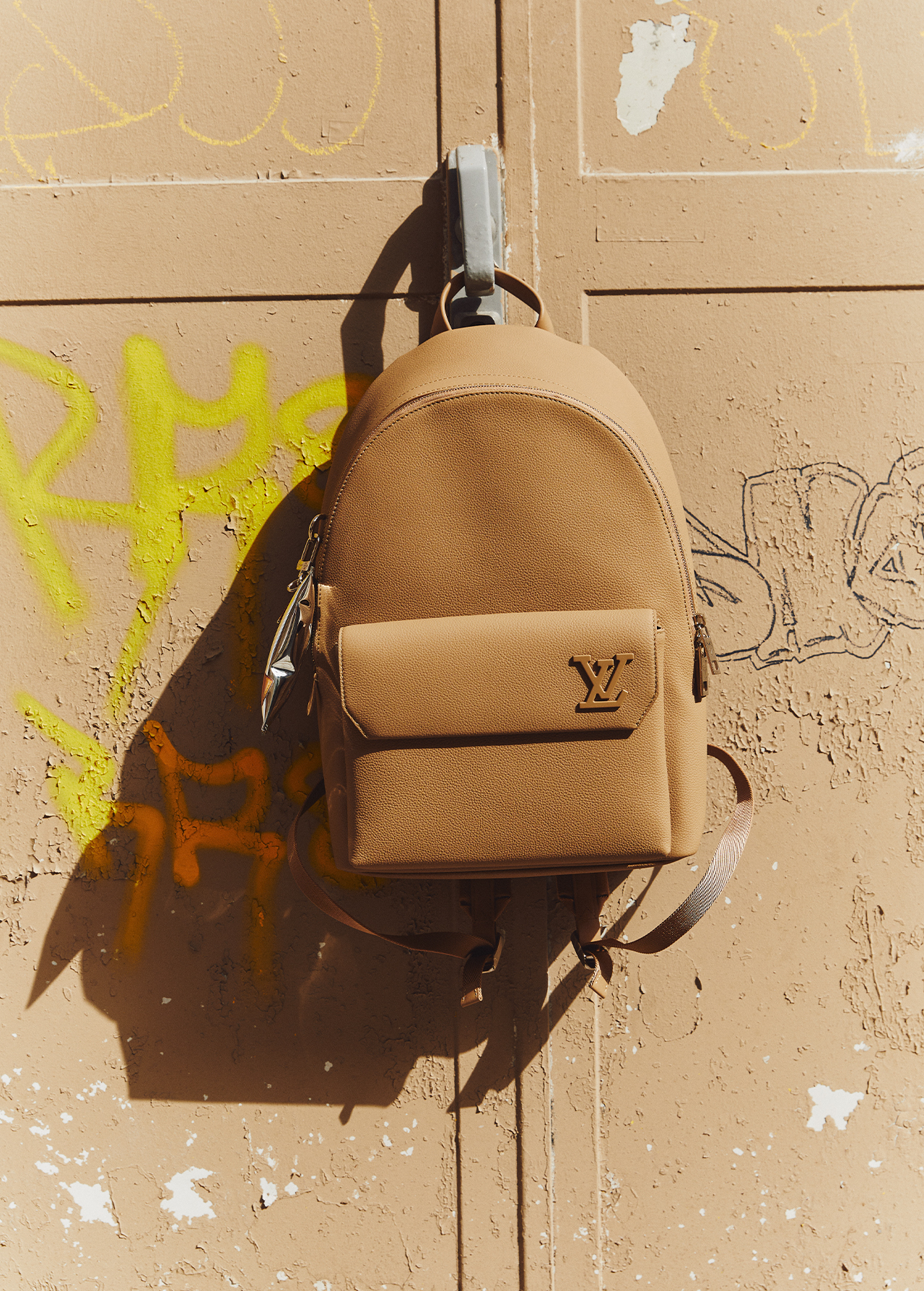 Tan leather backpack with a small front pocket and the logo, hangs from a hook on a textured wall with colorful graffiti in the background.
