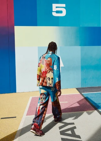 Fashionable adult wearing a multicolor face-print suit, styled hair, and sneakers, posing in front of an abstract painted wall.