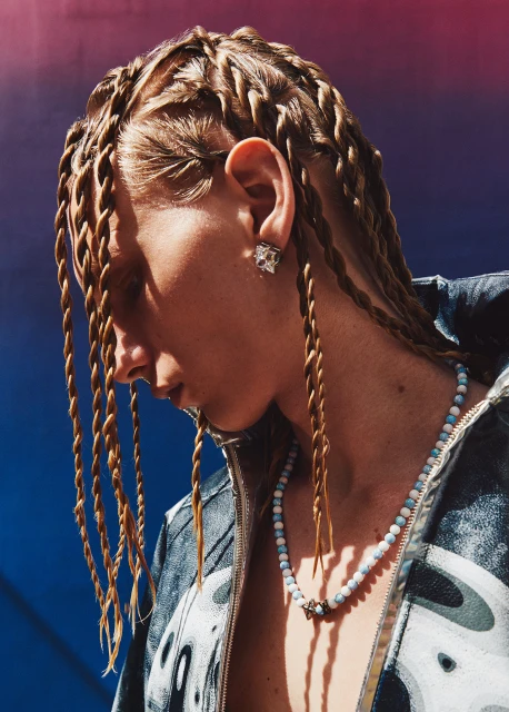 Close-up of a young adult with tan skin wearing tan braids, a blue beaded necklace, and silver earring posing against a blue and red background.