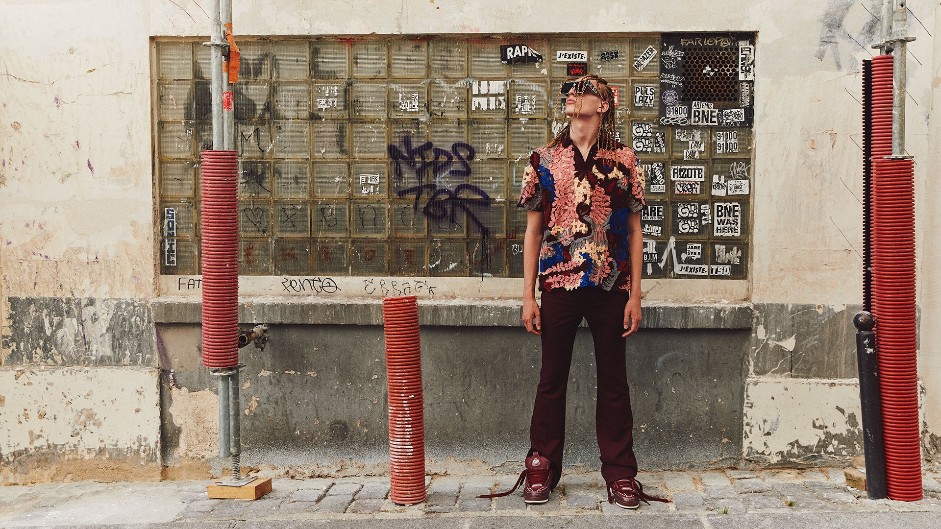 Man in patterned shirt and maroon pants stands against a graffiti wall in an outdoor urban setting, gazing upwards in a fashion-forward pose.