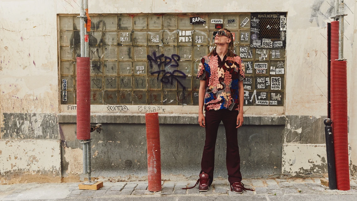 Man in patterned shirt and maroon pants stands against a graffiti wall in an outdoor urban setting, gazing upwards in a fashion-forward pose.