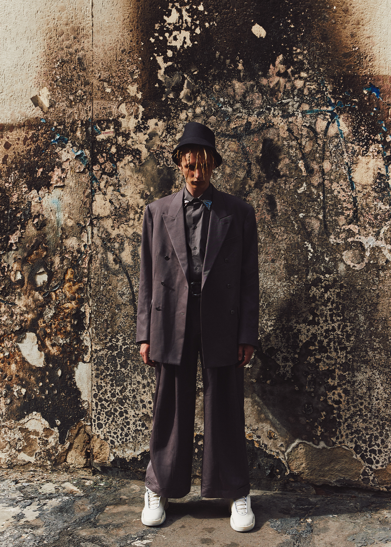 Young adult poses in an oversized grey suit with a black hat and white sneakers against a distressed concrete wall in an urban environment.