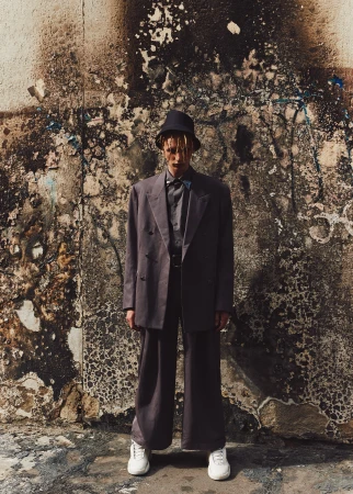 Young adult poses in an oversized grey suit with a black hat and white sneakers against a distressed concrete wall in an urban environment.