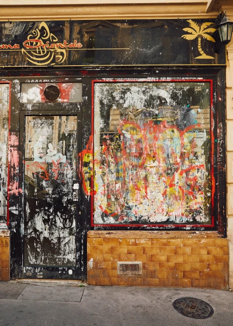 Exterior of weathered storefront with graffiti, boarded-up windows, and peeling paint in urban setting during daylight hours.