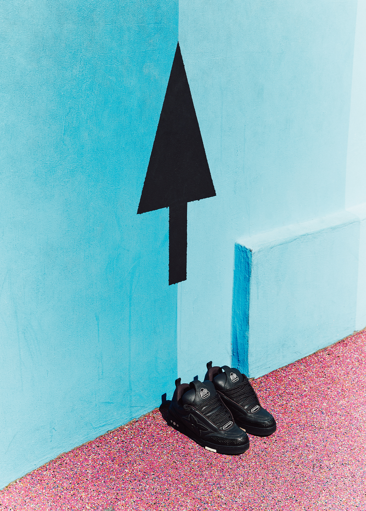 Two black shoes on a pink textured surface, set against a light blue wall with a painted black arrow pointing upwards.