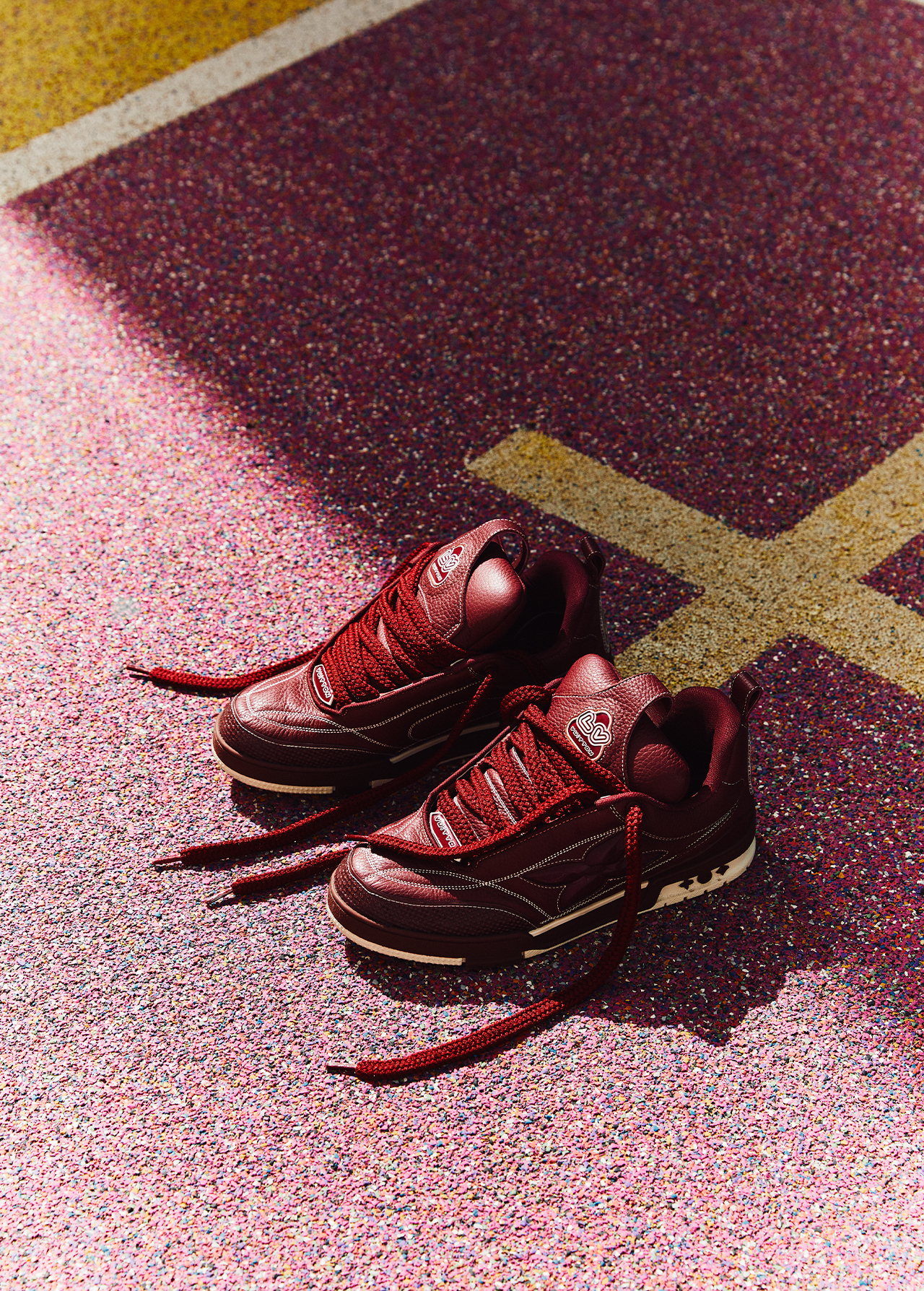 Close-up of burgundy sneakers on a pink and maroon speckled surface with graphic yellow lines. Sneakers are angled slightly, laces untied.
