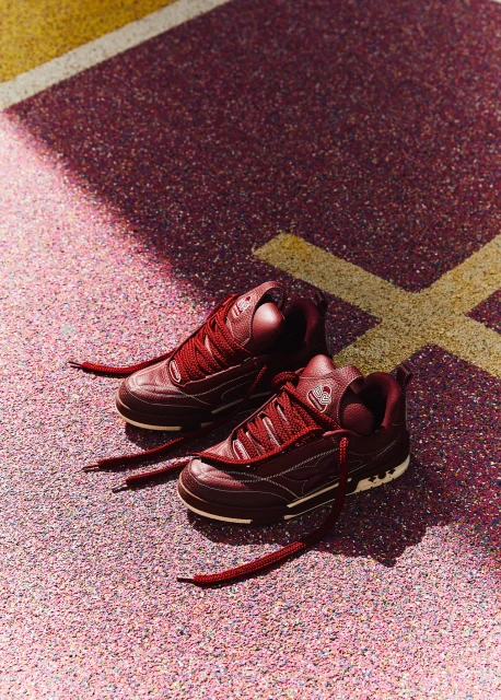 Close-up of burgundy sneakers on a pink and maroon speckled surface with graphic yellow lines. Sneakers are angled slightly, laces untied.