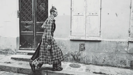 Model in a plaid trench coat walking past a weathered building exterior in a black and white editorial image