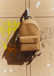 Tan leather backpack hanging on a weathered metal door with yellow graffiti in harsh sunlight.