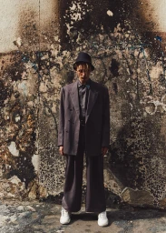 Full-length portrait of a man wearing an oversized grey suit, bucket hat, and white sneakers standing against a weathered wall