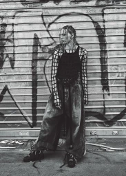 Model with braided hair wearing a net shirt and wide leg trousers standing in front of a graffitied metal shutter