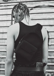 Monochrome portrait of a person with braided hair wearing a black tank top and crossbody bag against a metal shutter door.