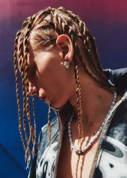 Close-up profile of a model with braided blonde hair wearing a patterned jacket, beaded necklace, and earrings against a gradient background.