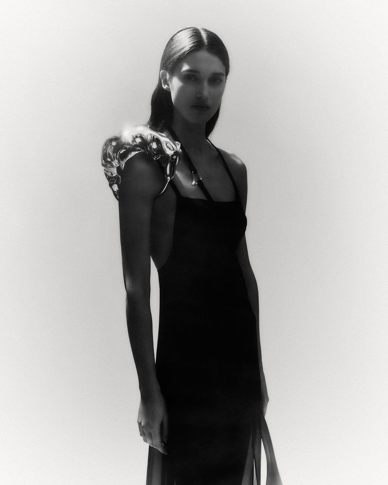 Model wearing a dark dress with a metallic sculptural shoulder piece against a white seamless background