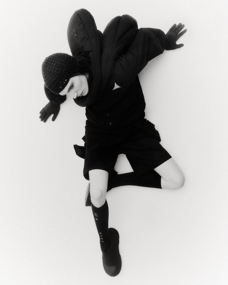 Fashion shot of a person in black knit cap, padded coat, shorts, high socks and shoes against a white backdrop in a studio. High angle.