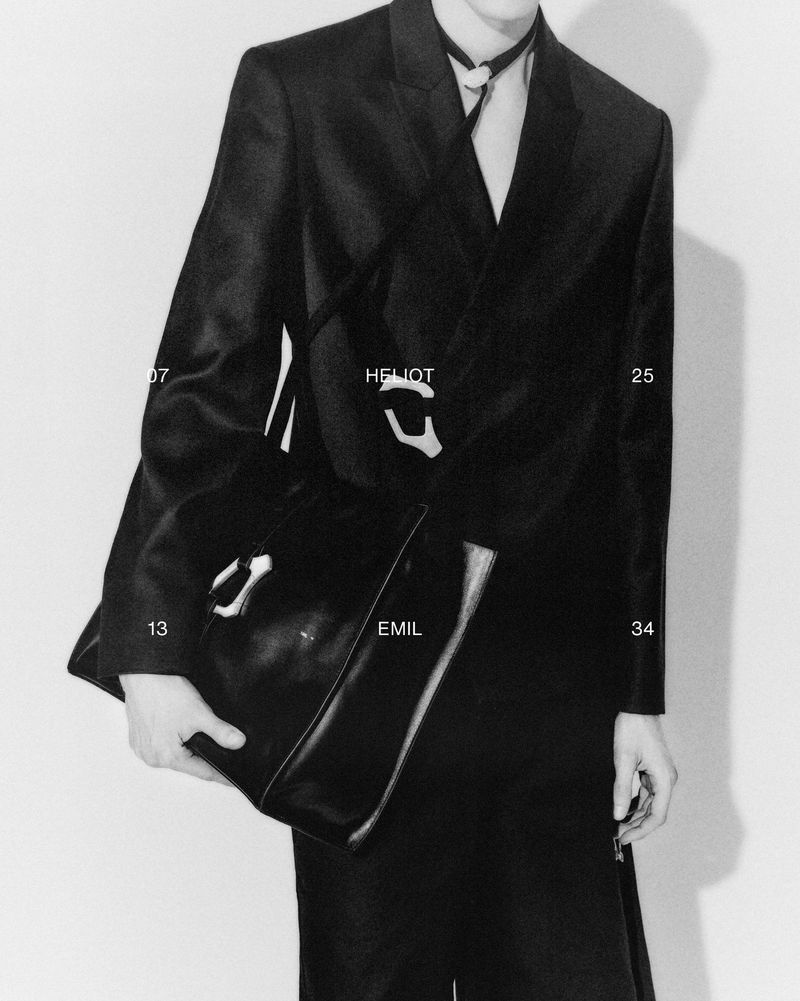 Elegant adult male model displays a black leather satchel with a statement clasp, wearing a dark suit in a minimalist studio setting.