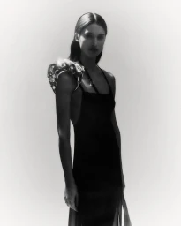 Model wearing a dark dress with a metallic sculptural shoulder piece against a white seamless background