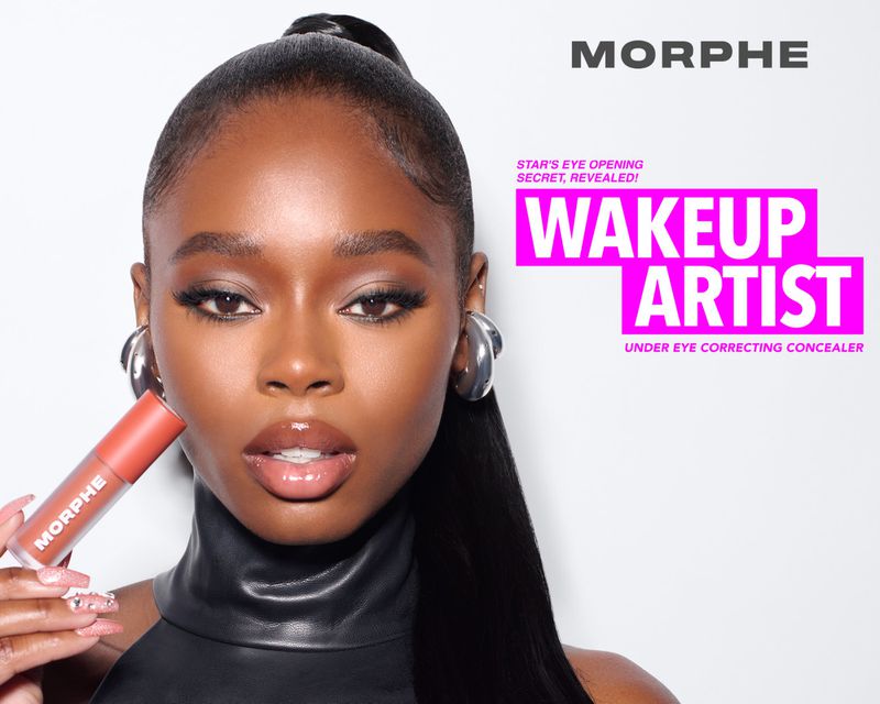 An adult female model with brown skin poses in a beauty shot holding Morphe makeup while wearing a black turtleneck and hoop earrings.