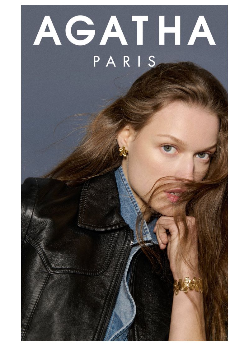 Blonde model wearing a black leather jacket with gold floral jewelry including earrings and a cuff bracelet, posing in front of gray background.