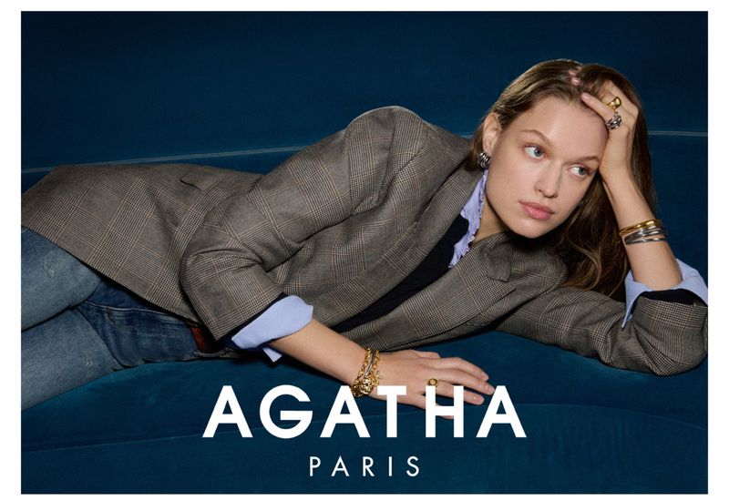 Woman poses wearing blazer and jewellery including rings and bracelets while reclining on a blue velvet surface, advertising AGATHA PARIS.