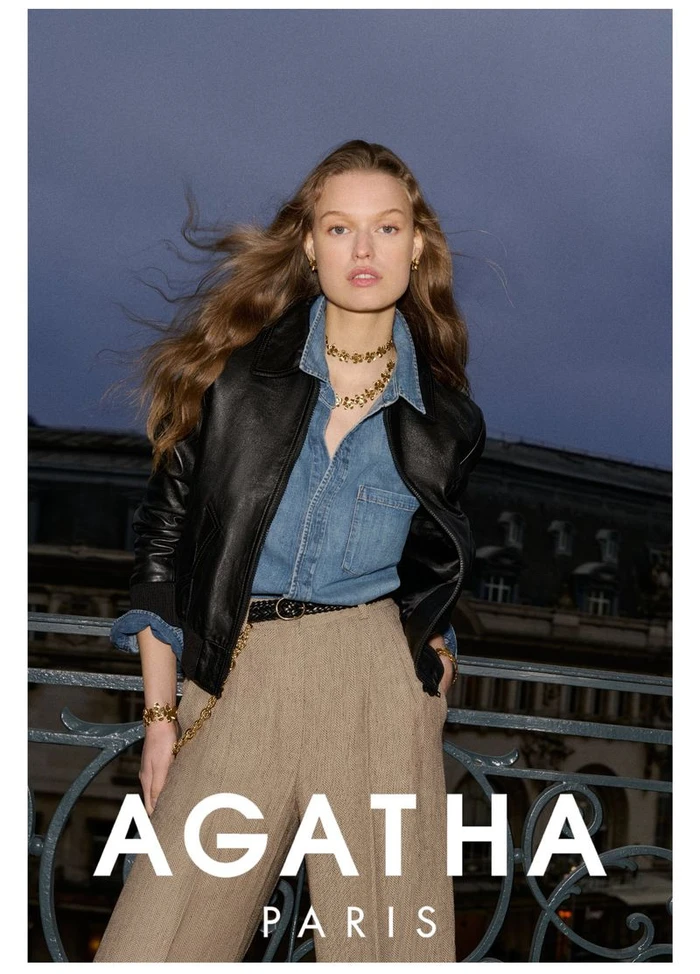 Model wearing a black leather jacket and denim shirt leaning against an ornate balcony railing outdoors at dusk.