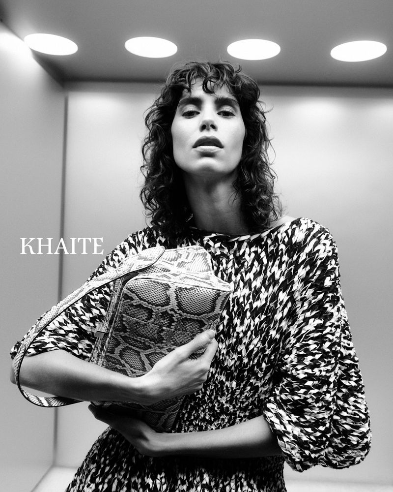 Black and white shot of a woman in patterned dress holding a snakeskin purse, standing indoors under recessed lighting.