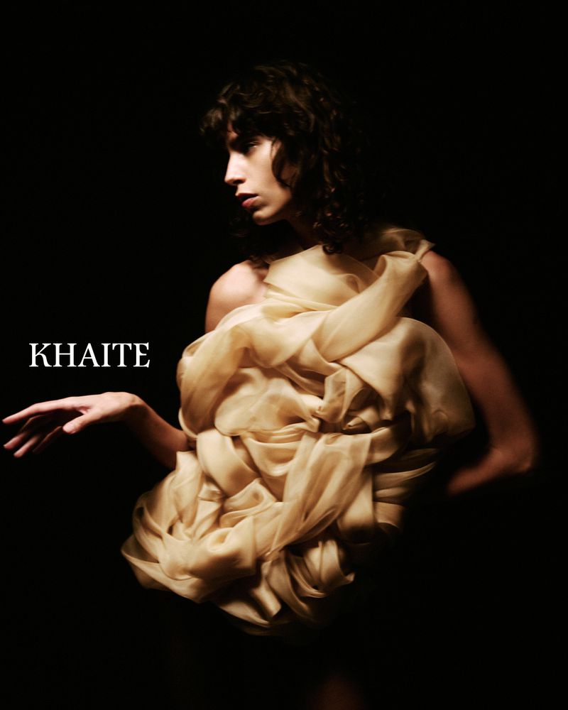 Woman draped in flowing beige fabric posing against a dark background in a studio fashion shot. The image includes the text 'KHAITE'.