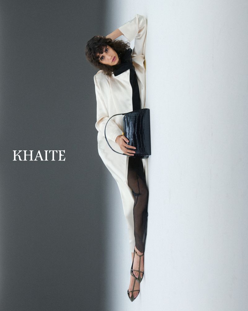 Model Mica Argañaraz wearing a white coat and black handbag against a split grey and white studio background for Khaite S/S 2025.