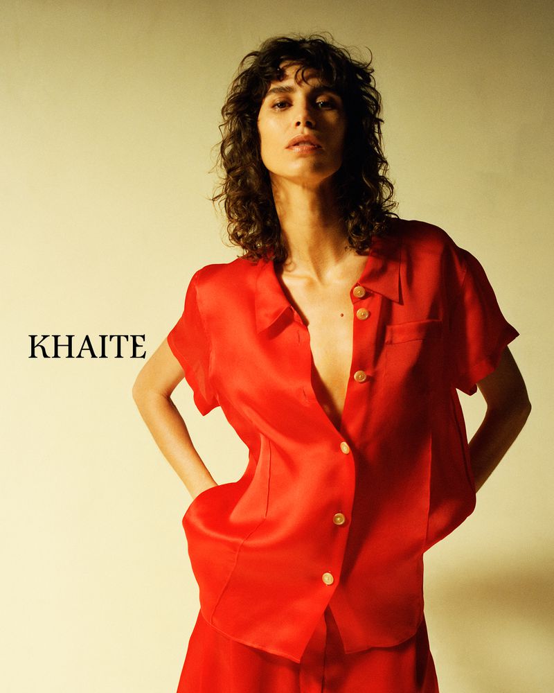 An adult female poses indoors wearing a red silk short-sleeve button-up shirt and a matching skirt against a pale yellow background.