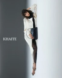 Model Mica Argañaraz wearing a white coat and black handbag against a split grey and white studio background for Khaite S/S 2025.