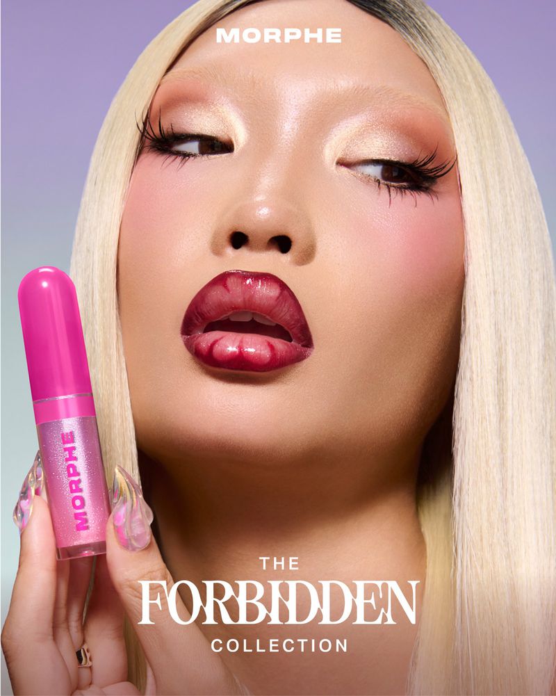 Close-up beauty shot of an adult female model with blonde hair, bright eyeshadow and red lips holding a tube of Morphe lip gloss.