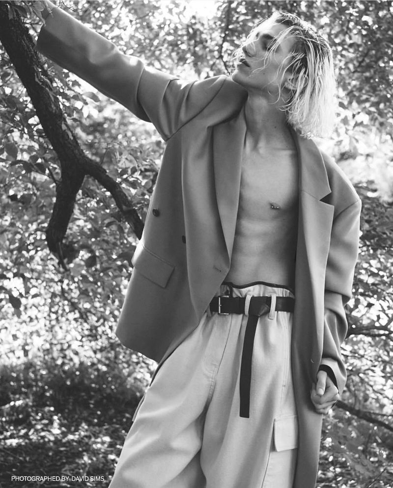 Black and white portrait of a young adult man wearing an open blazer and pants, posing outdoors among trees and foliage.