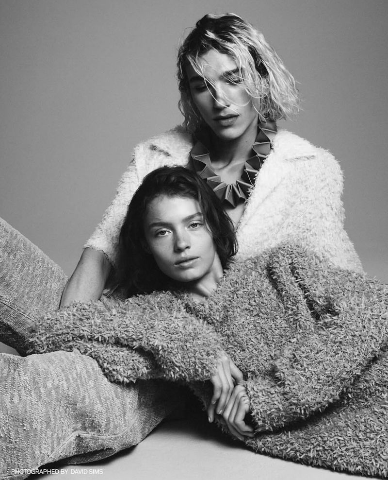 Black and white portrait of two models, one seated and one behind with short hair, wearing textured clothing against a neutral background.