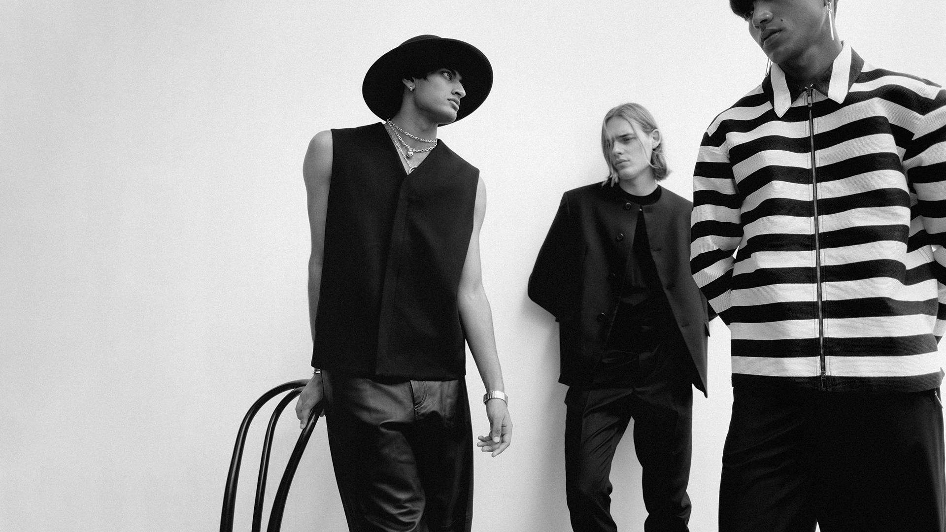 Three fashion models pose indoors against a plain white wall wearing stylish black and white striped clothing. A modern chair.