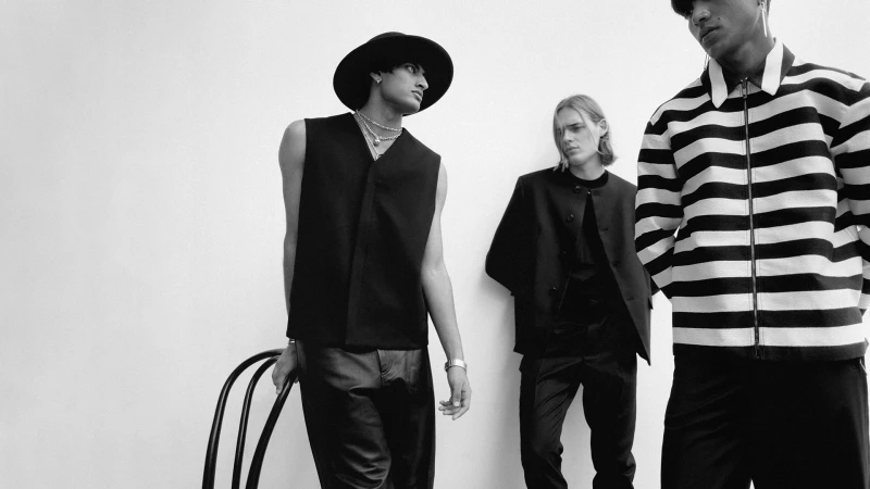 Three fashion models pose indoors against a plain white wall wearing stylish black and white striped clothing. A modern chair.