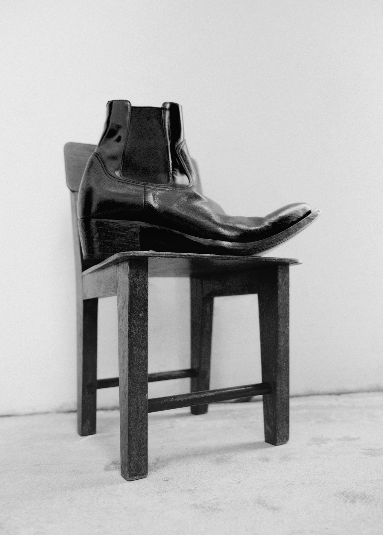 Black leather boot placed as the primary subject on a vintage-style wooden chair in a minimalist indoor setting against a white wall.